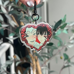 K&ouml;nnte beinhalten: Ein herzf&ouml;rmiger Acryl-Schl&uuml;sselanh&auml;nger mit zwei Anime-Charakteren. Ein Charakter hat wei&szlig;es Haar und der andere hat schwarzes Haar. Der Schl&uuml;sselanh&auml;nger hat einen roten Zierrand und einen schwarzen Verschluss.