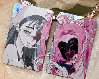 Puede incluir: Dos tarjetas coleccionables holográficas rectangulares con ilustraciones de estilo anime. La tarjeta de la izquierda muestra un personaje de pelo oscuro con pañuelo. La de la derecha muestra un personaje de pelo rosa y máscara de corazón.