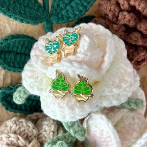 May include: A pair of gold-tone earrings with green enamel monstera leaf designs. The earrings are displayed on a white crocheted surface.