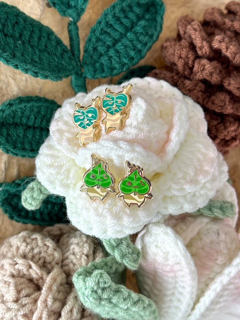 May include: A pair of gold-tone earrings with green enamel monstera leaf designs. The earrings are displayed on a white crocheted surface.