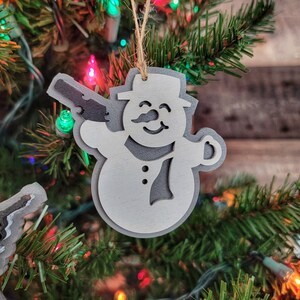 May include: White wooden snowman ornament with a gun in its hand. The ornament is hanging on a Christmas tree with red and green lights.