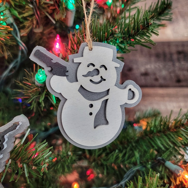 May include: White wooden snowman ornament with a gun in its hand. The ornament is hanging on a Christmas tree with red and green lights.