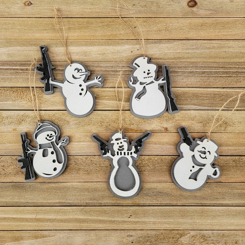 May include: Six white snowman ornaments with black guns and scarves. The ornaments are hanging from twine on a wooden background.