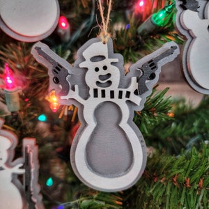 May include: A white snowman ornament with a black hat and scarf, holding two toy guns in each hand. The ornament is hanging on a Christmas tree with red and green lights.