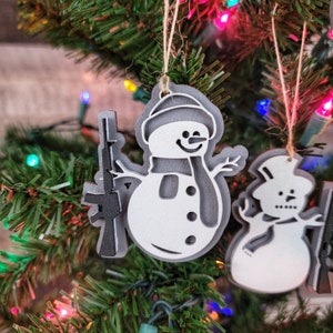 May include: Two white painted wooden ornaments shaped like snowmen. One snowman is holding a black and gray painted wooden rifle. Both snowmen have black painted eyes, mouths, and buttons. The snowmen are hanging on a Christmas tree with red, green, and white lights.