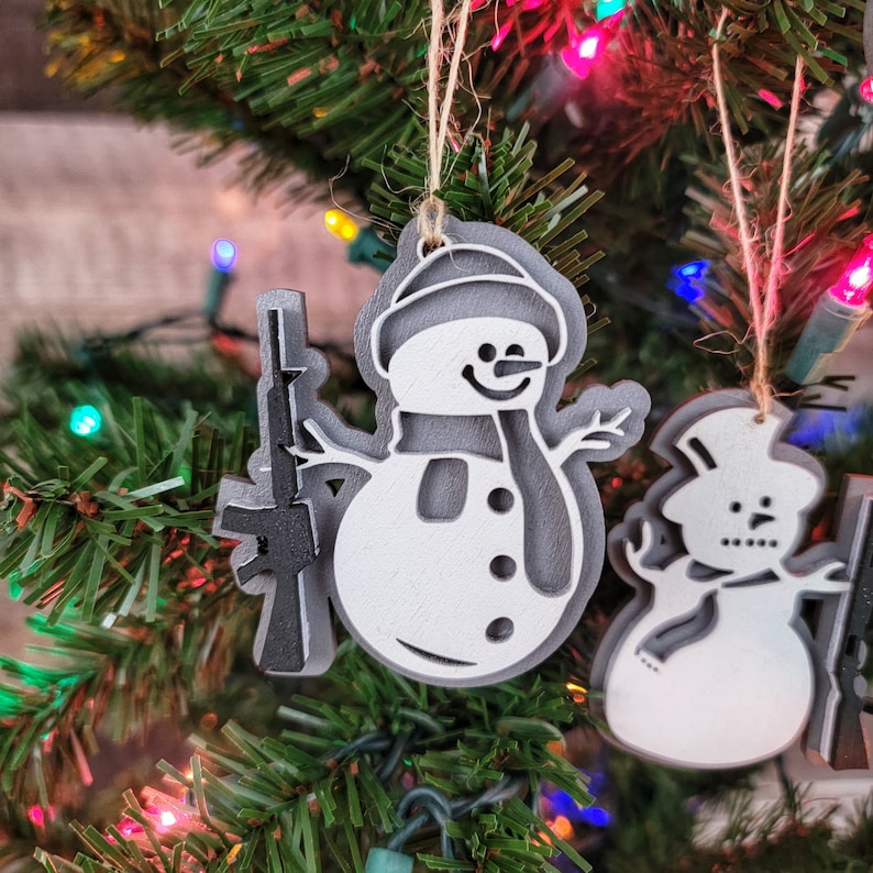 May include: Two white painted wooden ornaments shaped like snowmen. One snowman is holding a black and gray painted wooden rifle. Both snowmen have black painted eyes, mouths, and buttons. The snowmen are hanging on a Christmas tree with red, green, and white lights.