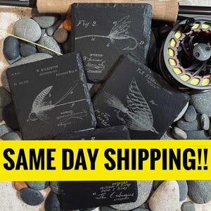 May include: Four black slate coasters with engraved illustrations of vintage fly fishing lures. The coasters feature detailed drawings of the lures, along with the text "Fly Fishing Lure" and "Fig. 2". The coasters are surrounded by small gray rocks.