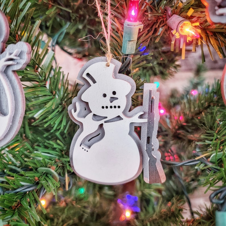 May include: White wooden snowman ornament with a gray scarf and a rifle. The ornament is hanging on a Christmas tree with colorful lights.