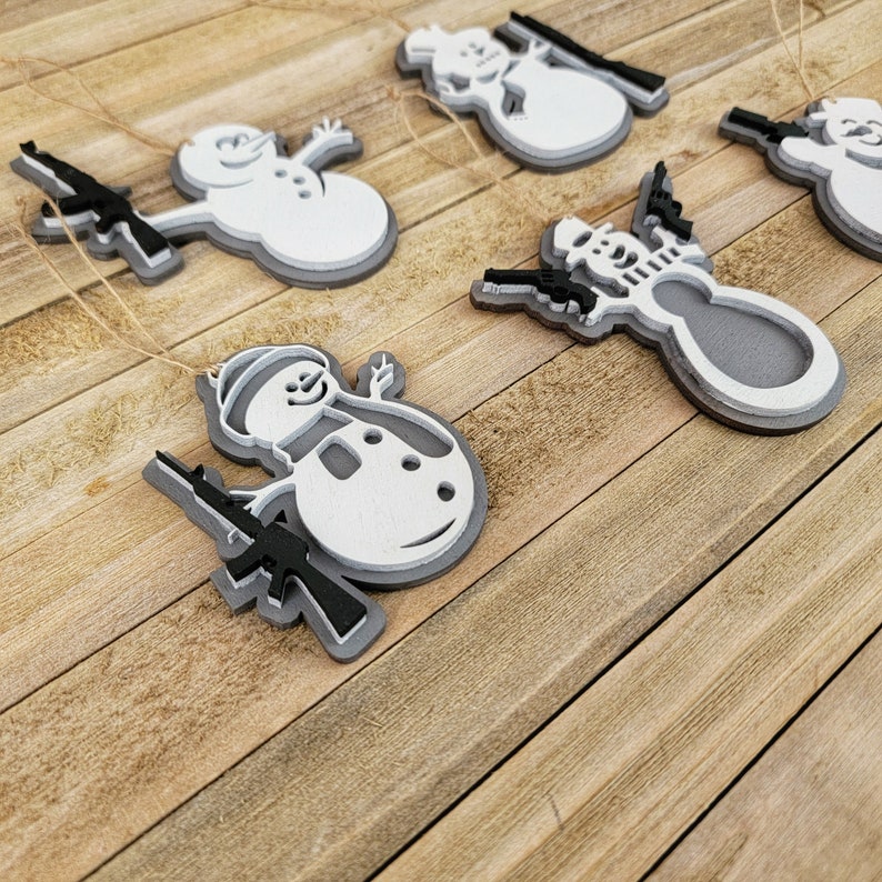 May include: Five white and gray snowman ornaments with black guns. The snowmen are holding the guns in their arms and are smiling. The ornaments are hanging from a string.