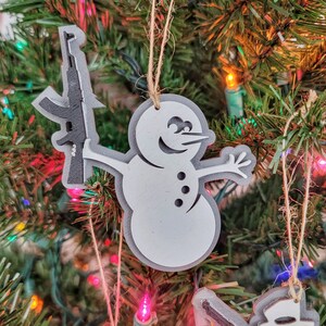 May include: A white snowman Christmas ornament holding a black rifle. The ornament is made of wood and has a rustic look.