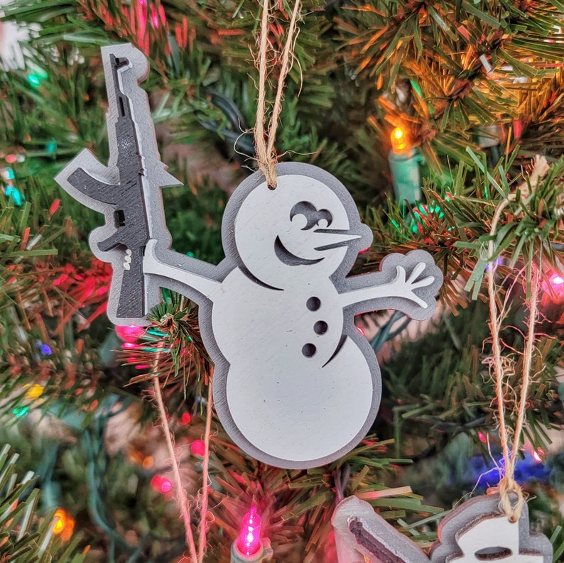 May include: A white snowman Christmas ornament holding a black rifle. The ornament is made of wood and has a rustic look.