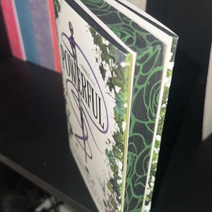 Könnte beinhalten: Ein Taschenbuch mit dem Titel "Powerful" mit einem grünen und schwarzen floralen Design auf dem Cover. Das Buch befindet sich in einem schwarzen Regal mit anderen Büchern dahinter.