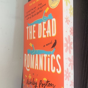 Könnte beinhalten: Ein rotes Taschenbuch mit dem Titel "The Dead Romantics" von Ashley Poston. Der Umschlag zeigt eine stilisierte Illustration von zwei Personen, die liegen, mit einem Vogel, der auf einem Ast darüber sitzt. Das Buch ist ein New York Times Bestseller und ein Good Morning America Book Club Pick.