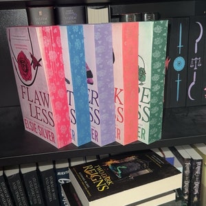 Könnte beinhalten: Ein Bücherregal mit einer Vielzahl von Büchern, darunter ein Stapel Bücher mit dem Titel "Two Dark Reigns" von Kendare Blake. Die Bücher im oberen Regal haben Titel wie "System Divine" und "Forever Rains".