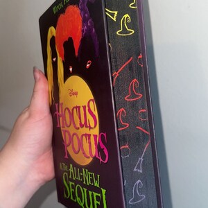 Peut inclure: Un livre relié en carton rigide de couleur violette et noire avec le titre "Hocus Pocus and the All-New Sequel" en lettres vertes et roses. La couverture présente une silhouette des trois sœurs Sanderson du film, avec le texte "Witch, Please." en haut.