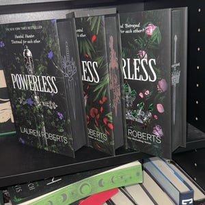 Könnte beinhalten: Auf einem Regal sind mehrere Hardcover-Bücher zu sehen, darunter "Powerless" von Lauren Roberts, mit einem dunklen Einband, der ein Schwert und florale Muster zeigt. Weitere Bücher mit ähnlichen Einbänden sind sichtbar.