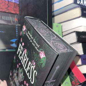Könnte beinhalten: Ein schwarzes Hardcover-Buch mit dem Titel "FEARLESS" von Lauren Roberts, mit rosa Rosen und einem Schwert-Design. Das Buch liegt auf einem Regal mit anderen Büchern.