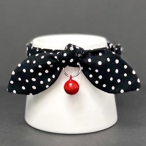 May include: Black and white polka dot fabric bow tie with a red bell charm. The bow tie is tied around a white ceramic base.