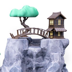 May include: A miniature zen garden scene featuring a waterfall cascading over gray rock formations. A small wooden bridge leads to a traditional Japanese house with lit windows. A green-leafed tree sits above the bridge.