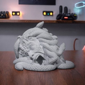 May include: A white, sculpted bust of Medusa, the Greek mythological figure, with snakes for hair. The bust is made of a smooth, white material and is a detailed representation of the mythical creature.