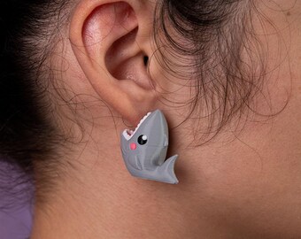 May include: A pair of gray shark earrings with a cute cartoon face and open mouth. The earrings are worn in a person's ear.