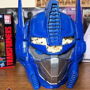 May include: A blue plastic Optimus Prime head with a silver faceplate. The head is filled with popcorn.
