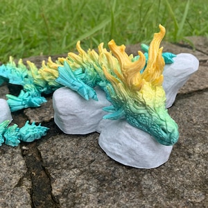May include: A detailed, colorful dragon figurine in shades of teal, green, yellow, and gold. The dragon is posed on a gray stone surface, with its head resting on a rock. The dragon's body is segmented, and the details are intricate.