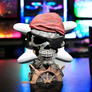 May include: A skull figurine with a red bandana, black eye patch, and a ship's wheel base. The skull is gray with a wide grin. Two white game controllers are on either side of the skull.