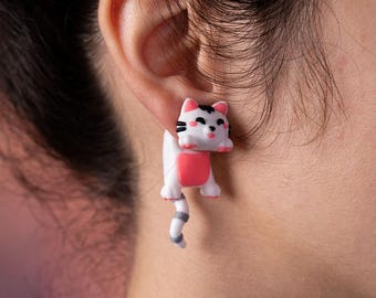 Peut inclure: Boucles d'oreilles fantaisie en forme de chat blanc avec des détails roses et noirs. La queue du chat pend, créant un effet ludique. Ces boucles d'oreilles sont un ajout amusant à toute collection de bijoux.