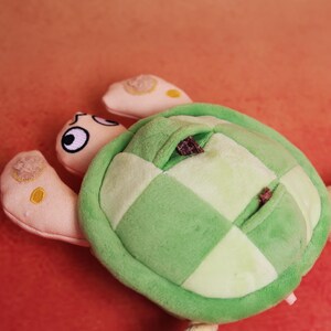 May include: A plush turtle toy with a green and white checkered shell, light green trim, and three beige flippers with yellow accents. The turtle's head is beige with black and white eyes. The toy is on an orange background.