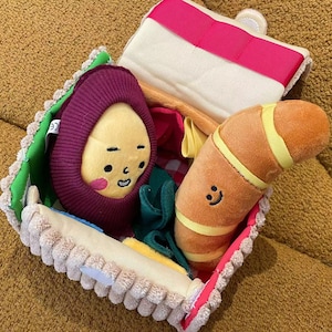 Könnte beinhalten: Ein Plüsch-Spielzeug-Essensset in einem sandwichförmigen Behälter. Es enthält eine lächelnde Avocado, ein Croissant und andere farbenfrohe Stoffstücke. Der Behälter hat eine beige, grüne und rosa Umrandung.