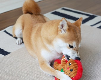 May include: A Shiba Inu dog playing with a red and brown gingerbread man plush toy. The dog is lying on a white and black striped rug.