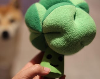 May include: A plush green broccoli dog toy with a smiling face. The toy is held in a hand, with a blurred dog in the background. The broccoli head is a lighter green than the stem.