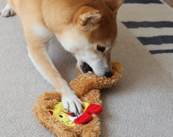 May include: A brown and white dog playing with a brown plush toy shaped like a bear. The toy has a yellow face with a red nose and a red bow.