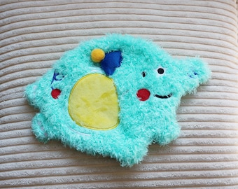 May include: A turquoise blue plush toy shaped like a cartoon character with a yellow circle and a blue and yellow star on top. The toy has a red circle for a nose and a black eye.