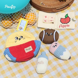 Puede incluir: Colección de juguetes de peluche para perros, que incluye un juguete con forma de perro amarillo y rojo, un balón de fútbol americano, un lápiz y un biberón. Los juguetes están colocados sobre una superficie a cuadros amarillos. La palabra "Pawfoo" es visible.