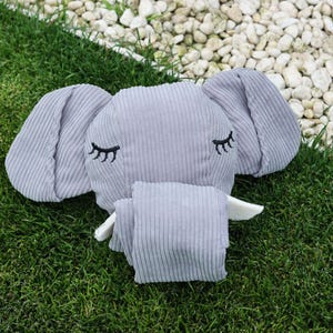 May include: A gray corduroy elephant-shaped plush toy with embroidered black eyelashes. The elephant has large, floppy ears and a small trunk. The toy is lying on green grass with white stones in the background.