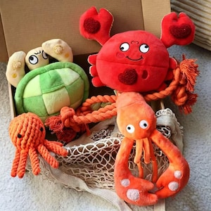 Puede incluir: Un conjunto de juguetes de peluche de criaturas marinas que incluye una tortuga verde y crema, un cangrejo rojo brillante con pinzas texturizadas y un pequeño pulpo naranja hecho de cuerda anudada, junto a un gran peluche con forma de langosta.
