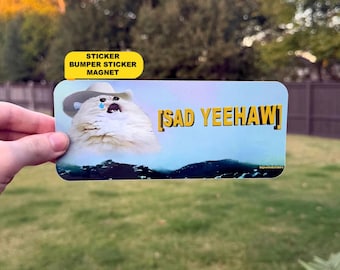 Puede incluir: Una pegatina o imán rectangular que muestra un gato blanco y esponjoso con un sombrero de vaquero y una lágrima azul en la cara. El texto SAD YEEHAW está impreso en letras amarillas sobre un cielo azul claro y un paisaje oscuro.