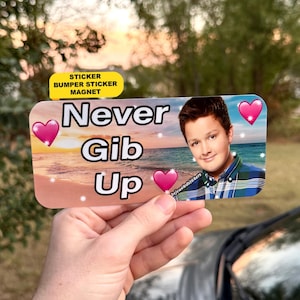 May include: A rectangular bumper sticker magnet featuring a sunset beach background, the text Never Gib Up in black letters, three pink hearts, and a photographic portrait of a person wearing a blue and green plaid button-down shirt.