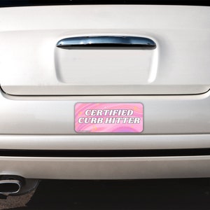 May include: A rectangular bumper sticker with a pink and purple swirled background featuring small yellow star accents. The text CERTIFIED CURB HITTER is printed in bold white capital letters with a dark outline centered on the sticker surface.