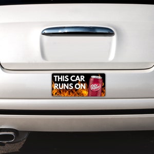 May include: A rectangular bumper sticker attached to the rear of a white car. The sticker features a black background with orange flames, a red Dr Pepper soda can, and the white text that reads: THIS CAR RUNS ON.