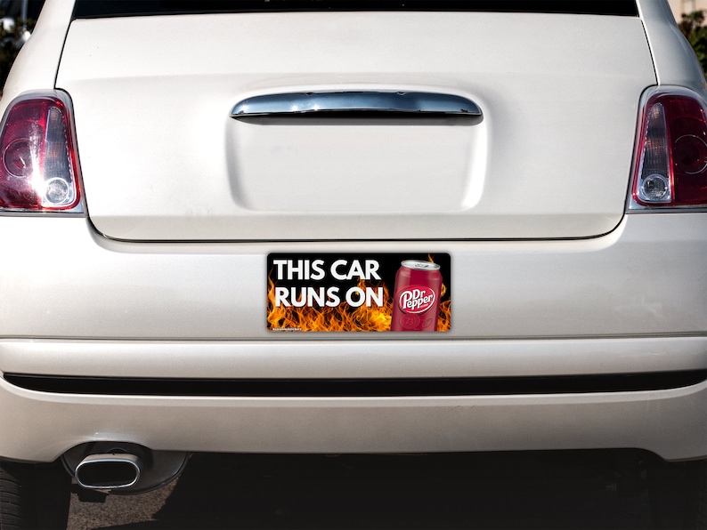 May include: A rectangular bumper sticker attached to the rear of a white car. The sticker features a black background with orange flames, a red Dr Pepper soda can, and the white text that reads: THIS CAR RUNS ON.
