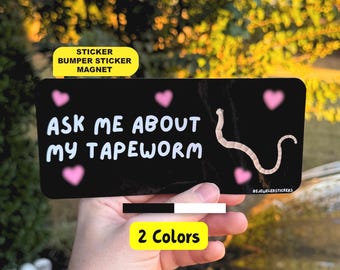 May include: A rectangular black bumper sticker or magnet featuring the white text ASK ME ABOUT MY TAPEWORM, decorated with five small pink hearts and a graphic illustration of a segmented tapeworm on the right side.