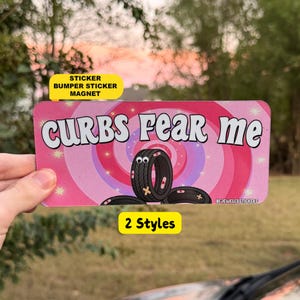 May include: A rectangular bumper sticker or magnet featuring a pink and purple spiral background with white stars. The text CURBS FEAR ME is printed in large white letters above a cartoon tyre with googly eyes and adhesive bandages.
