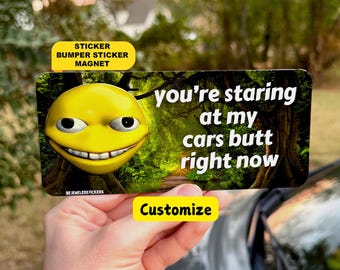 May include: A rectangular bumper sticker magnet featuring a yellow 3D-style smiley face with wide eyes and a toothy grin. The background shows a forest path. The text reads: you're staring at my cars butt right now.