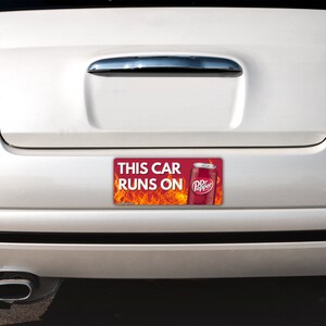 May include: A rectangular bumper sticker attached to the back of a white car. The sticker features a red background with orange flames, a graphic of a Dr Pepper soda can, and the white text that reads THIS CAR RUNS ON Dr Pepper.