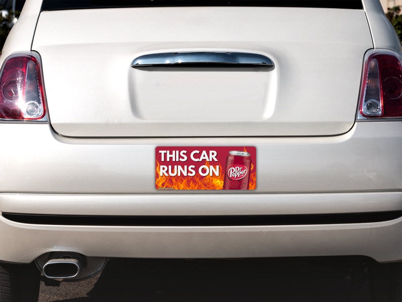 May include: A rectangular bumper sticker attached to the back of a white car. The sticker features a red background with orange flames, a graphic of a Dr Pepper soda can, and the white text that reads THIS CAR RUNS ON Dr Pepper.