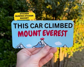 Może przedstawiać: Prostokątna jasnoniebieska naklejka z czarnym napisem THIS CAR CLIMBED i czerwonym napisem MOUNT EVEREST. Poniżej tekstu znajduje się grafika ośnieżonego szczytu góry z małym czerwonym samochodem i białymi chmurami.