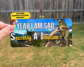 Może przedstawiać: Prostokątna naklejka na zderzak przedstawiająca zielonego dinozaura w tropikalnym krajobrazie. Napis głosi YEAH I AM SAD Secretly A Dinosaur białymi i żółtymi literami. Naklejka ma żółtą zakładkę u góry z czarnym tekstem.